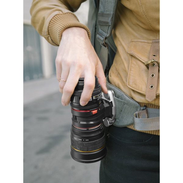 Peak-Design-Lens-Kit-Canon-6 Ulkona kaupungin kadulla ruskeaan takkiin pukeutunut henkilö käyttää Peak Design Lens Kit - Canon EF -objektiivisarjaa kiinnittääkseen kameran objektiivin turvallisesti vyöhönsä siihen tarkoitetulla pidikkeellä.