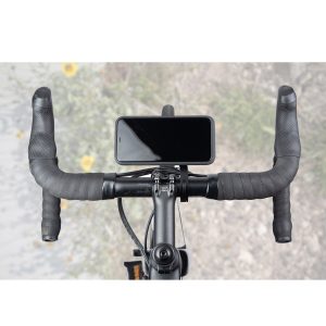 Ylhäältä alas näkymä mustasta polkupyörän ohjaustangosta, jossa on Peak Design Mobile Out Front Bike Mount -pyöräkiinnike, johon on kiinnitetty älypuhelin. Sumea tausta paljastaa kallioperän ja keltaisia kukkia.