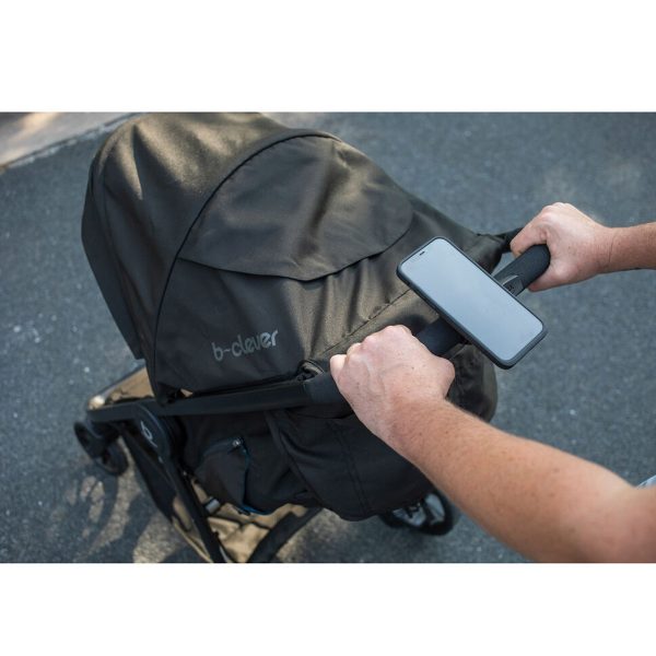Henkilö työntää mustia rattaita, joissa lukee "b-clever". Älypuhelin on kiinnitetty ohjaustankoon Peak Design Mobile Universal Bar Mount - Mustalla. Kohtaus tapahtuu ulkona päällystetyllä pinnalla.