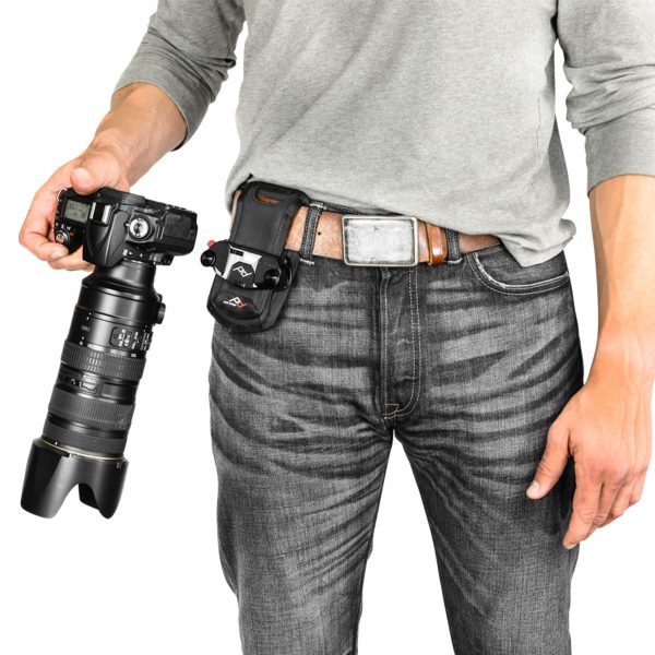 A man in a grey shirt and jeans is holding a camera with a large lens in his right hand. The Peak Design ProPad V2 camera case is attached to the left side of the belt at the waist.