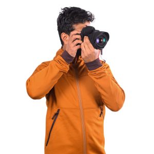 A person wearing an orange jacket is holding a Peak Design Shell Camera Cover - Small and taking a photo against a white background.