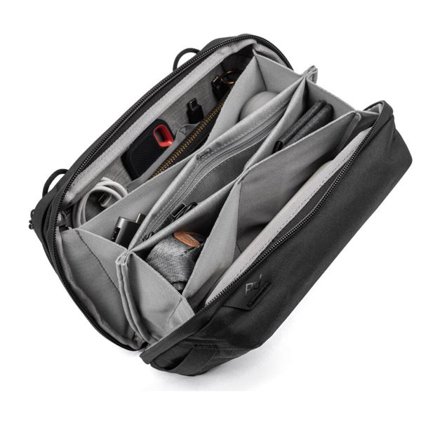 Peak Design Tech Pouch - Black on musta ja harmaa organisointilaite, jossa on useita lokeroita, joihin mahtuu siististi kaapeleita, kyniä, vetoketjullinen pussi ja elektronisia tarvikkeita. Avoimesta pussista näkyy kaikki sen järjestetty sisältö.