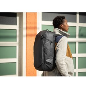 Ulkona oleva henkilö seisoo sivuttain rakennuksen edessä, hänellä on yllään harmaa ja ruskea fleecetakki ja hänellä on selässään musta Peak Design Travel Backpack 30L -kamerareppu.