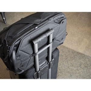 Peak Design Travel Backpack 45L -kamerareppu, musta on kiinnitetty tukevasti rullalaukun teleskooppikahvaan, joka lepää pilkullisella lattialla.