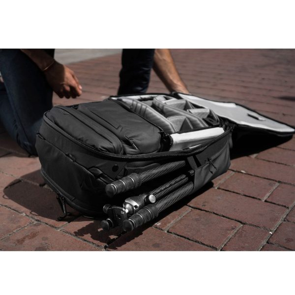 Henkilö polvistuu tiilipäällysteisellä jalkakäytävällä avoimen Peak Design Travel Backpack 45L -kamerareppu, musta, jonka sisälle mahtuu kameravarusteet ja sen ulkolokeroon osittain säilytetty jalusta.