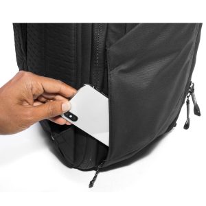 Käsi laittaa valkoisen älypuhelimen mustan Peak Design Travel Backpack 45L -kamerareppu -repun vetoketjulliseen sivutaskuun, jossa on pääosin valkoinen tausta.