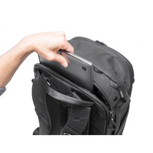 Käsi asettaa kannettavan tietokoneen mustan Peak Design Travel Backpack 45L -kamerareppu -repun etulokeroon valkoista taustaa vasten.