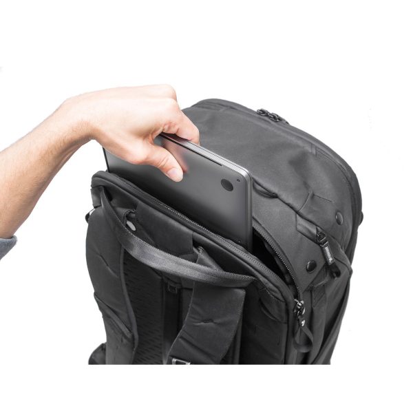 Käsi asettaa kannettavan tietokoneen mustan Peak Design Travel Backpack 45L -kamerareppu -repun etulokeroon valkoista taustaa vasten.