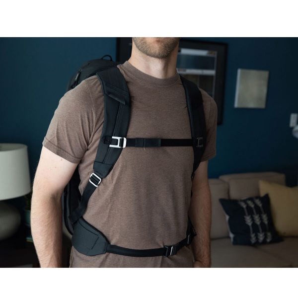 Ruskeaan t-paitaan pukeutunut henkilö, jolla on mukanaan Peak Design Travel Backpack 45L -kamerareppu, musta seisoo olohuoneessa, jossa on sohva, tyynyjä, pöytävalaisin ja seinätaidetta. Hänen kasvonsa ovat osittain kuvan ulkopuolella.