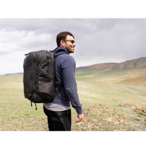 Aurinkolaseihin ja siniseen huppariin pukeutunut mies seisoo ruohoisella kukkulalla, jossa on pilvinen taivas ja kaukaiset vuoret, ja hänellä on Peak Design Travel Backpack 45L -kamerareppu, musta.