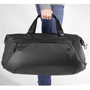 Sinisiin farkkuihin ja harmaaseen toppiin pukeutunut henkilö pitää Peak Design Travel Duffel 65L - Black -matkakassia kahvoista käsin valkoista taustaa vasten.