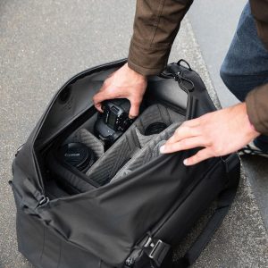 Henkilö asettaa Canon-kameran, ylimääräisiä objektiiveja ja tarvikkeita mustaan Peak Design Travel Duffel 65L -matkalaukkuun, jossa on järjestetyt lokerot, ja asettaa sen betonipinnalle.
