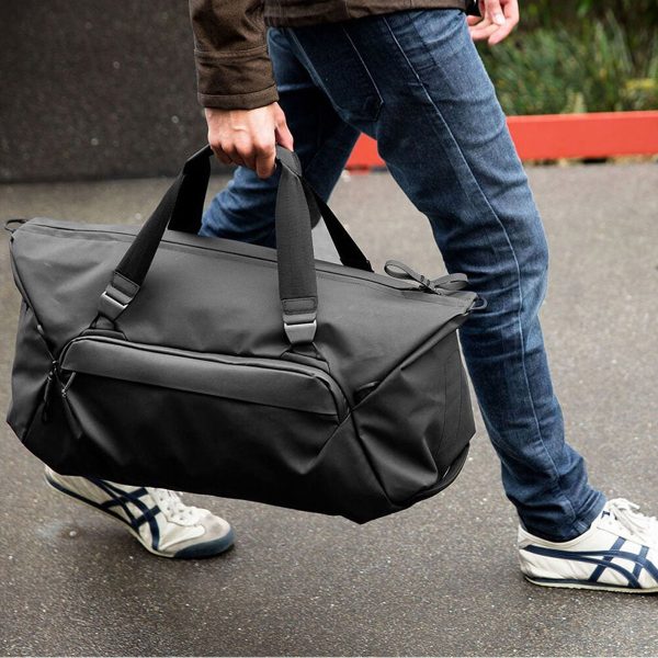 Ruskeaan takkiin, sinisiin farkkuihin ja valkoisiin lenkkareihin pukeutunut henkilö kävelee märällä jalkakäytävällä ja kantaa Peak Design Travel Duffel 65L - Black -matkalaukkua kahvoista.