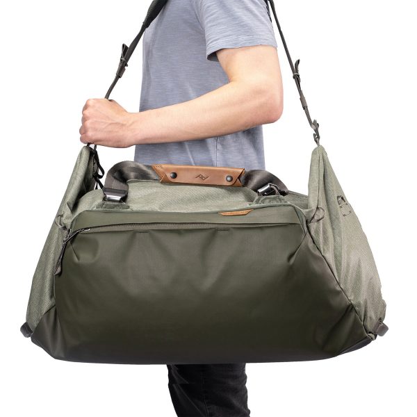 Vaaleansiniseen paitaan pukeutunut henkilö kantaa Peak Design Travel Duffel 65L - Sage -laukkua, joka on suuri vihreä ja harmaa laukku, jossa on nahka-aksentteja, useita lokeroita ja musta olkahihna.