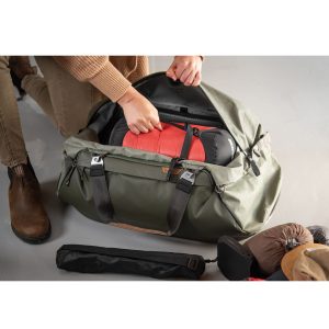 Ruskeapukuinen henkilö pakkaa punamustan makuupussin Peak Design Travel Duffel 65L - Sage -laukkuun. Erilaisia matkatarvikkeita lojuu lattialla vieressä.