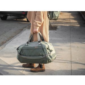 Henkilö, jolla on ruskehtava pusero, ruskeat housut ja ruskeat saappaat, seisoo jalkakäytävällä pitäen kädessään Peak Design Travel Duffel 65L - Sage -mallistoa. Hänen kasvonsa eivät näy. Taustalla on pysäköity auto ja reppu.