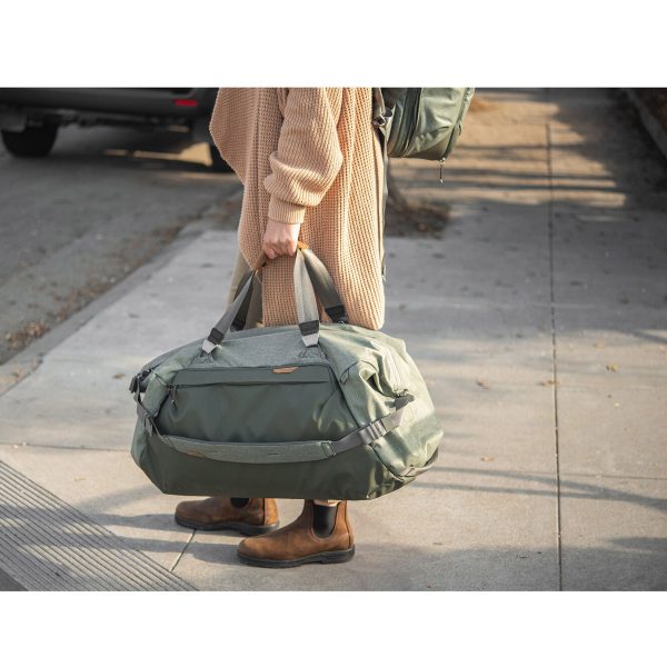 Henkilö, jolla on ruskehtava pusero, ruskeat housut ja ruskeat saappaat, seisoo jalkakäytävällä pitäen kädessään Peak Design Travel Duffel 65L - Sage -mallistoa. Hänen kasvonsa eivät näy. Taustalla on pysäköity auto ja reppu.