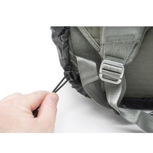 Lähikuva kädestä, joka vetää harmaan Peak Design Rain Fly - sadesuojan joustavaa kiristysnauhaa, reppuhihnat ja metallinen solki näkyvissä.