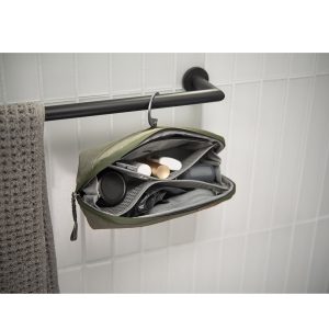 Peak-Design-Wash-Pouch-Small-Sage6 Peak Design Wash Pouch Small in Sage roikkuu auki mustasta pyyhetangosta harmaan pyyhkeen vieressä, ja sen sisällä on järjestettyjä hygieniatuotteita valkoista kaakeliseinää vasten.