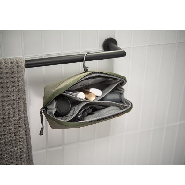 Peak-Design-Wash-Pouch-Small-Sage6 Peak Design Wash Pouch Small in Sage roikkuu auki mustasta pyyhetangosta harmaan pyyhkeen vieressä, ja sen sisällä on järjestettyjä hygieniatuotteita valkoista kaakeliseinää vasten.