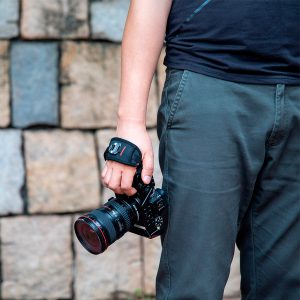 A person wearing a black shirt and grey trousers, holding a camera with a wide lens and wearing a SmallRig 2456 Hand Strap, stands in front of a stone wall.