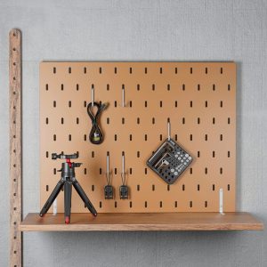 SmallRig-3184-6 A brown SmallRig 3184 Screw & Hex Key Storage Plate with coiled cable, two black adapters and hex keys is mounted on the light grey wall. A small tripod rests on a wooden shelf below.