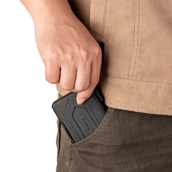 The person's hand is holding a black SmallRig 3192 memory card case, which is partially inserted into the side pocket of a pair of dark trousers with a beige jacket visible above.