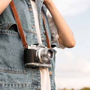 Henkilö, jolla on farkkuliivi, pitää kaulassaan Nikonin vintage-kameraa ja SmallRig 3485 Leather Camera Neck Strap -kamerahihnaa kaulan ympärillä sumeaa taivasta vasten.