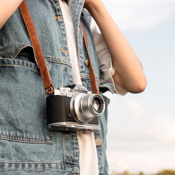 Henkilö, jolla on farkkuliivi, pitää kaulassaan Nikonin vintage-kameraa ja SmallRig 3485 Leather Camera Neck Strap -kamerahihnaa kaulan ympärillä sumeaa taivasta vasten.