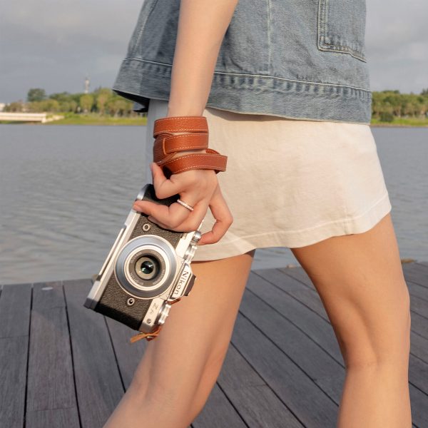 Valkoiseen hameeseen ja farkkutakkiin pukeutunut henkilö kävelee puisella laiturilla järven rannalla pitäen kameraa SmallRig 3485 Leather Camera Neck Strap -kamerahihnan kanssa. Taustalla näkyy puita ja sininen taivas.