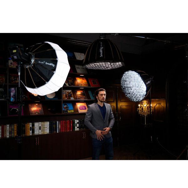 Harmaaseen bleiseriin pukeutunut mies seisoo hämärässä huoneessa kolmea SmallRig 3754 RA-L65 Lantern Softboxia vastapäätä. Hänen takanaan on kirjahyllyjä, joissa on värikkäitä kirjoja, ja koristeellinen seinävalaisin.