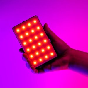 SmallRig-3808-6 Käsi pitelee SmallRig 3808 RGB Videolight RM120 -valaisinta, joka loistaa kirkkaan punaisilla ja keltaisilla LED-valoilla eloisan vaaleanpunaisen ja violetin värisävyisen taustan edessä.