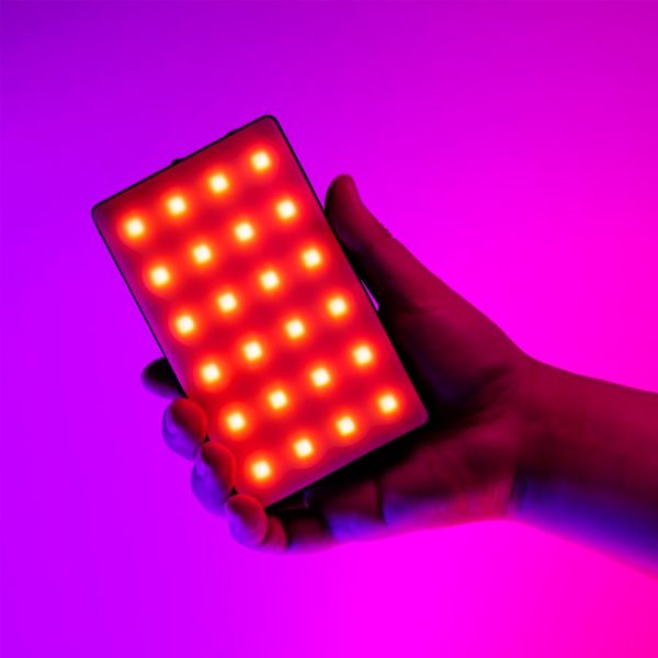 SmallRig-3808-6 Käsi pitelee SmallRig 3808 RGB Videolight RM120 -valaisinta, joka loistaa kirkkaan punaisilla ja keltaisilla LED-valoilla eloisan vaaleanpunaisen ja violetin värisävyisen taustan edessä.