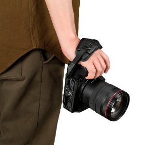 Ruskeisiin vaatteisiin pukeutunut henkilö pitää oikeassa kädessään ammattikäyttöön tarkoitettua kameraa, jossa on suuri objektiivi ja joka on kiinnitetty SmallRig 3848 Hand Strap Black Mamba-rannehihnalla, valkoista taustaa vasten.