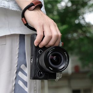 Henkilö, jolla on SmallRig 3926 Universal Wrist Strap Vintage -rannehihna, pitää kädessään mustaa kameraa, jossa on alaspäin suunnattu suuri linssi, ja taustalla on sumeaa vehreyttä ja rakennus.