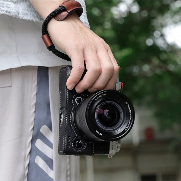 Henkilö, jolla on SmallRig 3926 Universal Wrist Strap Vintage -rannehihna, pitää kädessään mustaa kameraa, jossa on alaspäin suunnattu suuri linssi, ja taustalla on sumeaa vehreyttä ja rakennus.