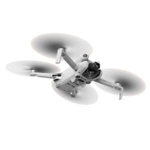 DJI Mini 4 Pro -lennokki, valkoinen nelikopteri, jossa on pyörivät potkurit, näkyy kesken lennon valkoista taustaa vasten. Etupuolen alapuolella on sen kamera ja anturit.
