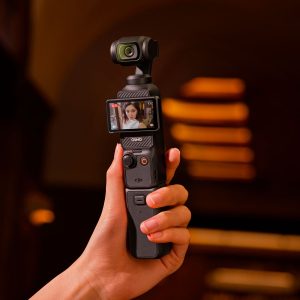 In her hand is a DJI Osmo Pocket 3 battery grip attached to a camera, whose rotating screen shows a woman's face against a warmly lit, blurred background in shades of orange and brown.