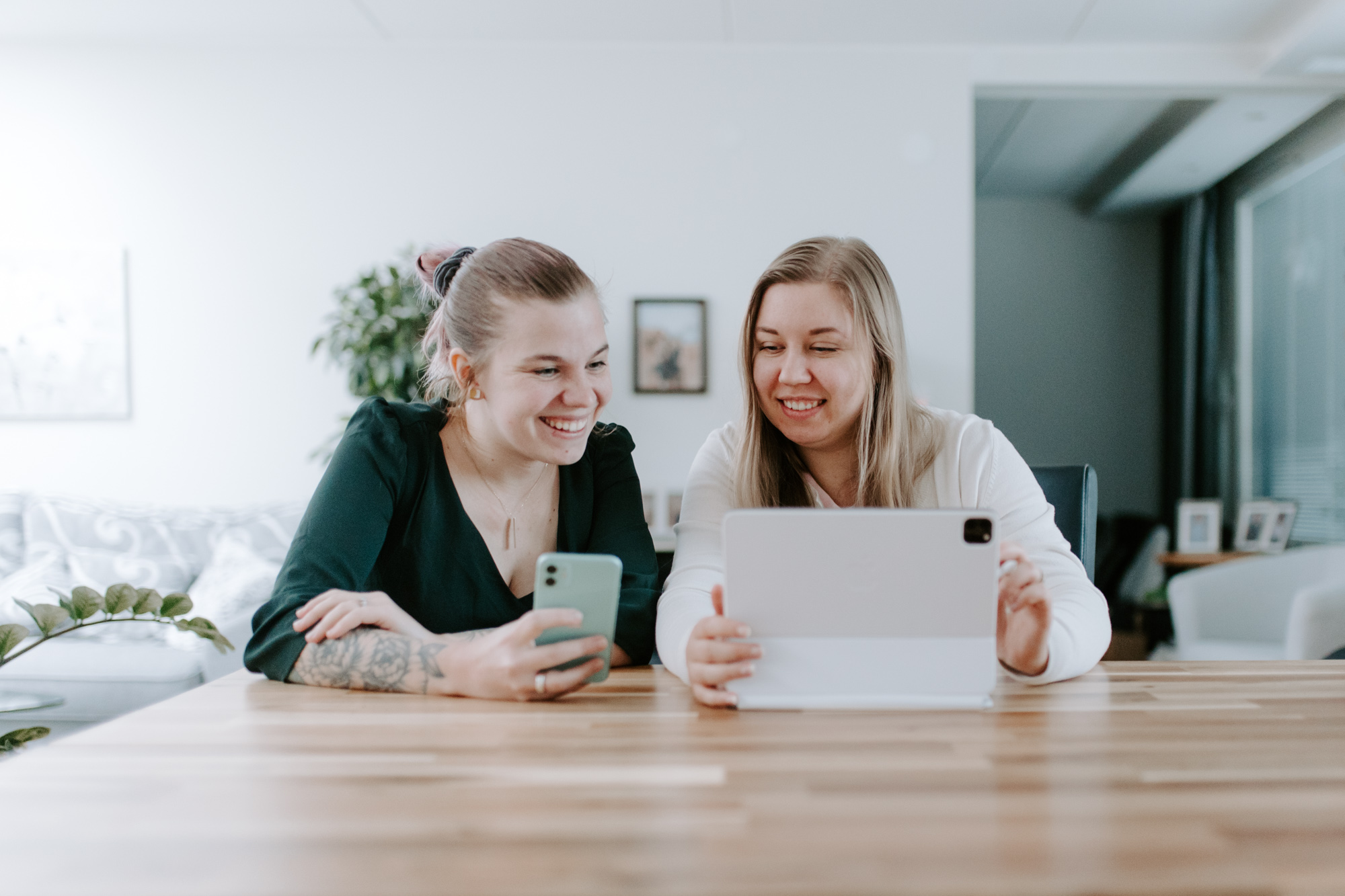 Kaksi naista istuu puupöydän ääressä hymyillen ja katselee tablettia ja älypuhelinta valoisassa ja modernissa sisätilassa.