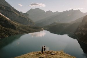 Two people stand on a grassy cliff overlooking a peaceful lake, surrounded by mountains in a clear sky, sunlight peeking through distant peaks.