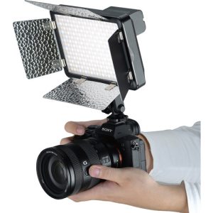 Henkilö pitää kädessään Sony-kameraa, jossa on suuri objektiivi ja suorakulmainen Godox Led LF308BI -videovalaisin, jonka päälle on asennettu heijastavat lato-ovet.
