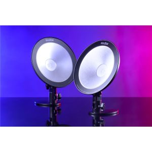 Kaksi Godox CL10 Ambient LED -valaisinta on sijoitettu vierekkäin jalustalle, ja ne hehkuvat sinistä ja vaaleanpunaista sävyä, ja niiden valo heijastuu kiiltävästä pinnasta.