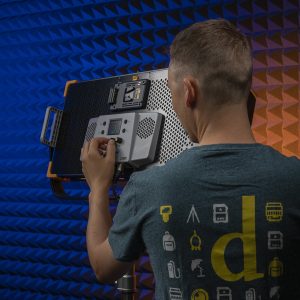 Henkilö, jolla on vihreä paita, jossa on teknisiä kuvakkeita, säätää Godox LED LD75R -studiovaloa, jossa on rei'itetty takapaneeli, seisoo sinisen ja oranssin akustisen vaahtomuovitaustan edessä.