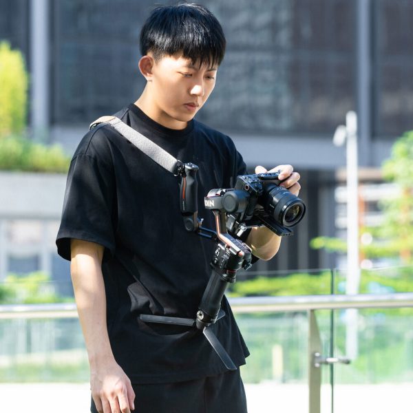 A black-shirted person uses the SmallRig 4118B Weight Reducing Shoulder Strap for DJI RS 3/RS 3 Pro/RS 2/RSC 2 with a camera outdoors amidst modern buildings and greenery.