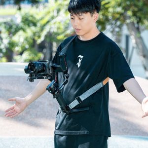 A person wearing a black t-shirt wears the SmallRig 4118B Weight Reducing Shoulder Strap for DJI RS 3/RS 3 Pro/RS 2/RSC 2 to support the camera's stabilizer while shooting outside on a sunny day with trees and greenery in the background.