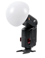 Godox Witstro Diffusor Ball, suuri valkoinen kupolimuotoinen kamerasalaman diffuusori, on kuvassa kiinnitettynä salamalaitteeseen ja asennettuna pieneen mustaan jalustaan valkoista taustaa vasten.