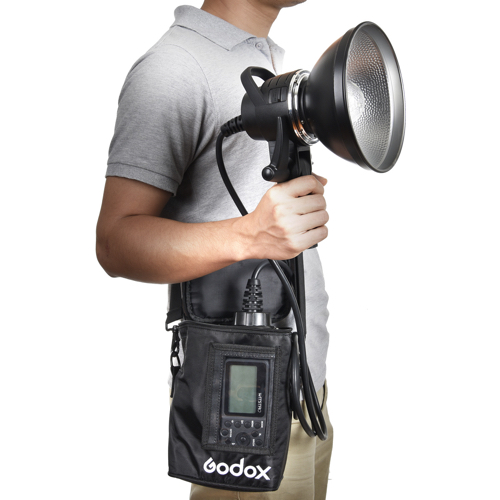 Harmaaseen poolopaitaan pukeutunut henkilö pitää kädessään Godox AD-H1200B Portable Flash Head Bowens -salamalaitetta, jossa on suuri heijastin ja joka on liitetty kaapelilla sen ohjausyksikköön, jota kannetaan mustassa olkalaukussa.