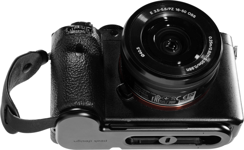 Musta Sony peilitön kamera, jossa on 16-50 mm:n objektiivi, on kuvassa ylhäältä alaspäin kiinnitettynä mustaan Peak Design Micro Clutch L-Plate -laattaan, jossa on rannehihna. Objektiivin yksityiskohdat ja kameran säätimet ovat näkyvissä.