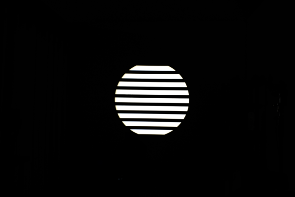 NANLITE GOBO Set 1 for PJ-FMM heijastaa kirkasta valkoista valoa vaakasuorilla säleillä varustetun pyöreän ikkunan läpi, jolloin pyöreän muodon sisällä olevat raidalliset kuviot näkyvät pimeässä huoneessa.