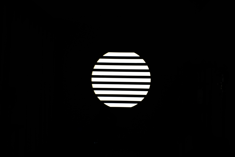 NANLITE GOBO Set 1 for PJ-FMM heijastaa kirkasta valkoista valoa vaakasuorilla säleillä varustetun pyöreän ikkunan läpi, jolloin pyöreän muodon sisällä olevat raidalliset kuviot näkyvät pimeässä huoneessa.